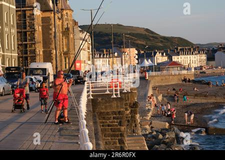 Mann in roten Shorts, Meeresangeln von der Meereswand bei Aberystwyth, an der walisischen Küste bei Sonnenuntergang. Mit einem weißen Geländer und Menschen, die sich unten sonnen. Stockfoto