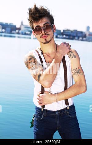 Sonnenbrille, Porträt und ein echter Mann auf dem Meer mit stilvollem Tattoo, Körperkunst und Mode. Punk, Ozean und trendiger Mann, der draußen in Spanien steht Stockfoto
