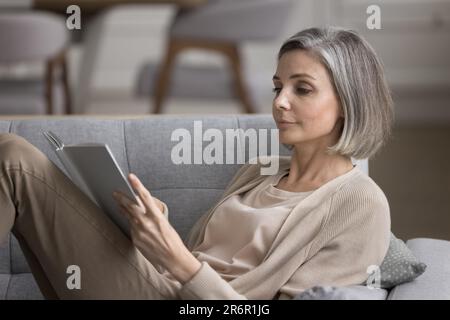 Eine fokussierte, grauhaarige Frau, die ein Bestseller-Buch liest Stockfoto