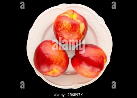 Drei organische Nektarinen mit einer weißen Platte auf schwarzem Hintergrund, Makro, Draufsicht. Stockfoto