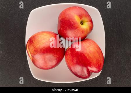 Drei organische Nektarinen mit einer weißen Platte auf schwarzem Hintergrund, Makro, Draufsicht. Stockfoto