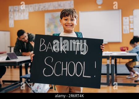 Aufgeregtes Kind, bereit für den ersten Tag der gemeinsamen Schulbildung und lebenslangen Lernens. Glücklicher Junge, der in einem Klassenzimmer steht, mit einem "Zurück zur Schule" -Schild. Elefanten Stockfoto