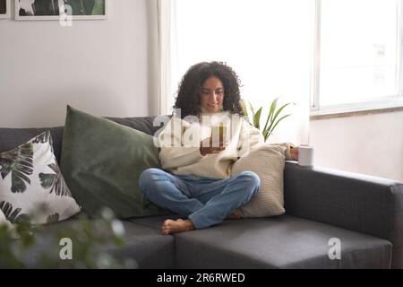 Lateinische glückliche junge Frau, die zu Hause auf dem Sofa mit Handy sitzt. Stockfoto
