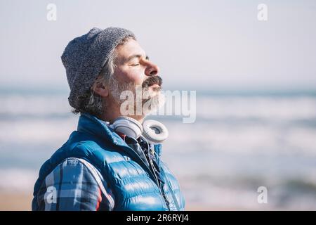 Ein fröhlicher junger Mann steht an einem windigen Strand und trägt Kopfhörer Stockfoto