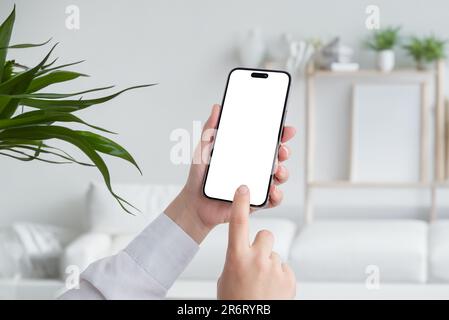 Junge Person, die ein modernes Mobiltelefon mit leerem weißen Bildschirm in der Hand hält. Wohnzimmereinrichtung im Hintergrund. Isolierter Bildschirm für Modellerstellung, App-Präsentation Stockfoto