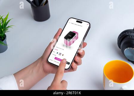 Smartwatch online mit Smartphone einkaufen. Person, die ein modernes Smartphone an einem Tisch im Haus hält, umgeben von einer Tasse Bleistifte, einer Zimmerpflanze und Stockfoto