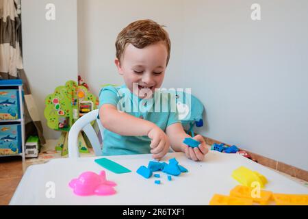 Der kleine Junge spielt enthusiastisch mit Plastizin, spielt zu Hause Teig auf dem weißen Tisch, das Kreativitätskonzept für Kinder Stockfoto