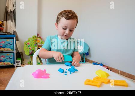 Der kleine Junge spielt enthusiastisch mit Plastizin, spielt zu Hause Teig auf dem weißen Tisch, das Kreativitätskonzept für Kinder Stockfoto