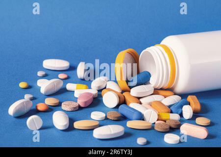 Viele Pillen sind aus einer Plastikflasche verschüttet worden. Mehrfarbige Pillen, die aus einer Plastikflasche auf blauem Hintergrund austreten. Eine Vielzahl von Pillen und Medikamenten. Stockfoto