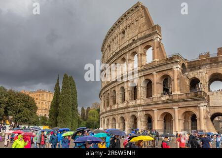 Besucher mit Sonnenschirmen bei starkem Regen und dunklem Himmel über dem Kolosseum, Rom, Italien Stockfoto