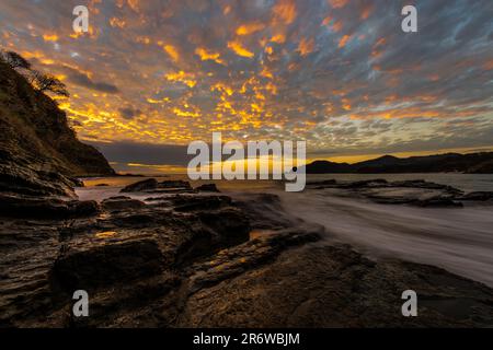 Lange Exposition Landschaftsfoto des pazifischen Ozeans, das über Felsen am Strand fließt, mit einem farbenfrohen Sonnenuntergang in der Nähe von San Juan del Sur in Nicaragua Stockfoto