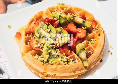 Große Waffel mit frischen Erdbeeren, Kiwischeiben, Pistazienteilen und Honig auf weißem Teller Stockfoto