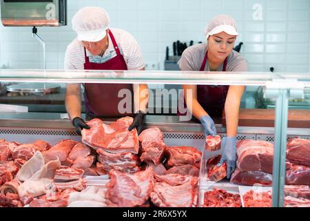 Ein älterer Verkäufer bereitet Schweinerippchen für den Kunden zu. Die Assistentin hat Rindfleisch ins Fenster gesteckt Stockfoto