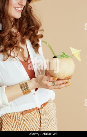 Strandurlaub. Lächelnde moderne Frau mittleren Alters in weißer Bluse und Shorts isoliert auf beigefarbenem Hintergrund mit Kokosnuss-Cocktail. Stockfoto