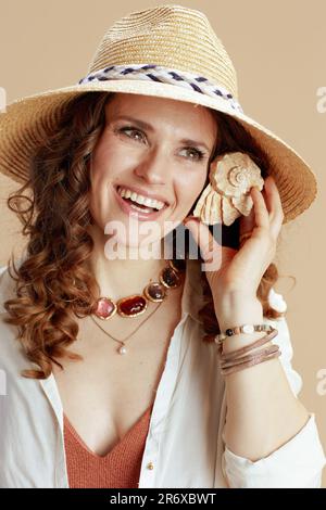 Strandurlaub. Lächelnde elegante Frau in weißer Bluse und Shorts auf beigefarbenem Hintergrund mit Strohhut und Muschelhören. Stockfoto