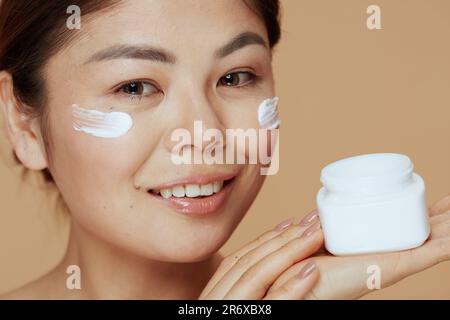 Porträt einer modernen asiatischen Frau mit Gesichtscremebecher und Gesichtscreme im Gesicht auf beigefarbenem Hintergrund. Stockfoto