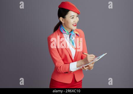 Lächelnde, stilvolle asiatische Stewardess in rotem Rock, Jacke und Hut Uniform mit Klemmbrett vor grauem Hintergrund. Stockfoto