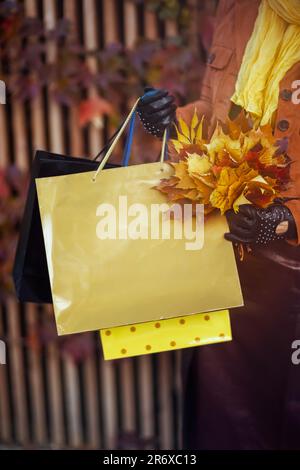 Hallo oktober. Nahaufnahme einer Frau mittleren Alters im orangefarbenen Trenchcoat mit Einkaufstaschen und herbstgelben Blättern in der Stadt. Stockfoto