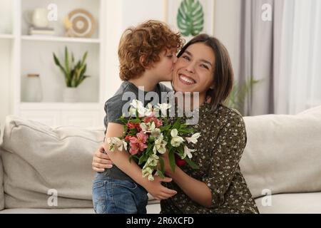 Der kleine Sohn küsst und gratuliert seiner mutter mit Muttertag zu Hause. Eine Frau mit Blumenstrauß Stockfoto