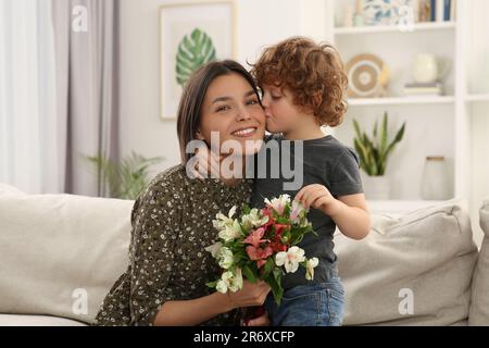 Der kleine Sohn küsst und gratuliert seiner mutter mit Muttertag zu Hause. Eine Frau mit Blumenstrauß Stockfoto