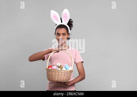 Glückliche afroamerikanische Frau mit Hasenohren Stirnband mit Korb aus Korb mit Ostereiern auf grauem Hintergrund Stockfoto
