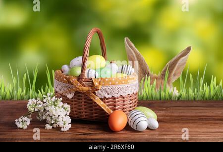 Bunny versteckt sich in grünem Gras neben einer Holzterrasse mit Korb voller Ostereier Stockfoto
