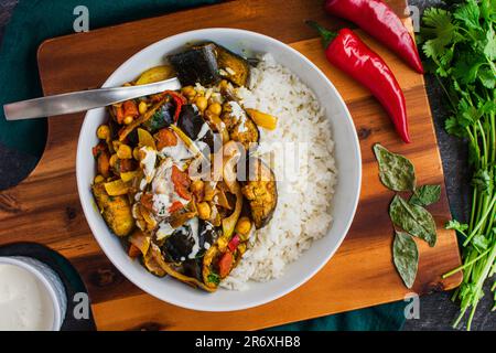 Vegane geröstete Auberginen- und Kichererbsen-Curry serviert auf Reis: Aubergine-Curry auf einer Schüssel weißen Reis und garniert mit pflanzlicher Sahne Stockfoto