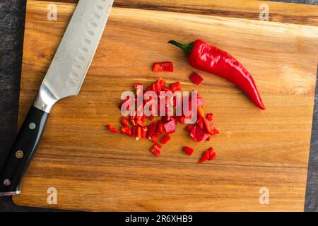 Gehackter roter Chili-Pfeffer auf einem Holzbrett: Ganzer roter chilipfeffer neben gewürfeltem chilipfeffer und Küchenmesser Stockfoto