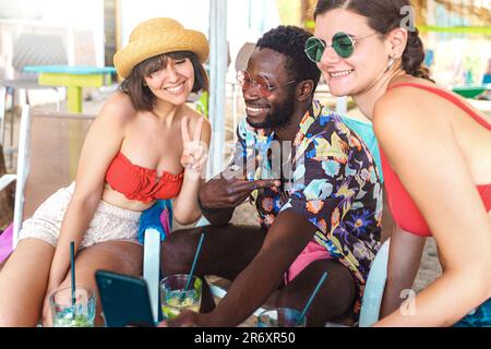 Ein unterhaltsamer Moment an der Strandbar, während ein junger Afrikaner mit seinem Smartphone für ein Selfie posiert. Seine beiden Freunde sind dabei, schauen auf das Telefon und SM Stockfoto