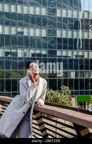 Vertikales Porträt einer jungen afrikanerin, die telefoniert. Sie hat Bürogebäude im Hintergrund Stockfoto