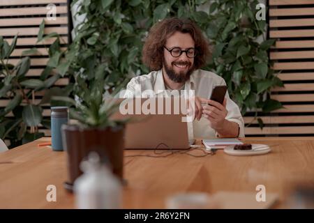 Junger, lächelnder Freiberufler, der an einem Laptop arbeitet und telefoniert, während er im Café sitzt. Konzept der Fernarbeit Stockfoto
