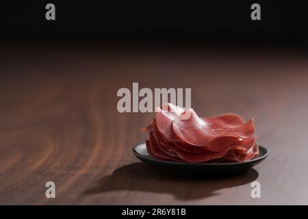 Untertasse mit diesen Bresaola-Scheiben auf Walnussholztisch, flacher Fokus Stockfoto