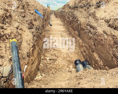 Bau einer neuen Nachbarschaft. Einen Graben graben, um Abwasserrohre zu verlegen. Abwassersysteme in Wohngebäuden. Breite schwarze Rohre im Boden. Stockfoto