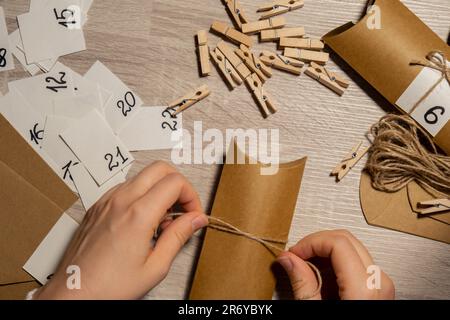 Eine nicht wiedererkennbare junge Frau klebt die Nummer auf die Handwerkstasche, wird mit einer Wäscheklammer befestigt. Weiblich macht Kraftpapier für hausgemachte Adventskalender mit eigenen Händen Schritt für Schritt selbst gemacht. Vorbereitung auf das weihnachtskonzept. Saisonale Aktivitäten für Kinder, Familienurlaub im Winter. Umweltfreundlich Geschenke. Öffnen Sie das Paket jeden Tag Stockfoto