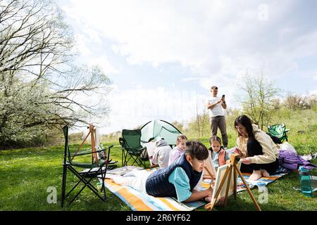 Glückliche junge Familie mit vier Kindern, die Spaß haben und sich im Freien auf Picknick-Decken-Malerei im Garden Spring Park amüsieren, Entspannung. Stockfoto