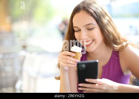 Eine glückliche Frau, die Eis isst und telefoniert, sitzt auf einer Barterrasse Stockfoto