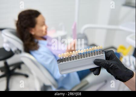 Vita-Waage. Zahnbleichführung. Zahnfarbtafel bei zahnärztlichen Händen über verschwommenen Patienten im zahnärztlichen Stuhl. Zahnheilkunde Stockfoto