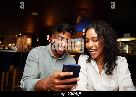 Ein junges, multiethnisches Paar in legerer Kleidung, das lacht und ein Smartphone in der Bar benutzt Stockfoto