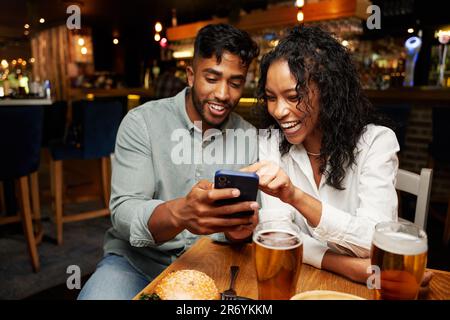 Ein junges, multiethnisches Paar in legerer Kleidung, das lächelt, während es im Restaurant auf dem Handy tippt Stockfoto