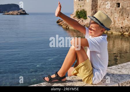 Ein stilvoller Teenager mit Strohhut sitzt am Meer und bedeckt das Sonnenlicht mit seiner Hand. Stockfoto