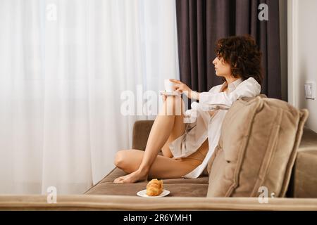 Die junge Frau sitzt im Profil auf dem Sofa, stellt eine Tasse Kaffee auf ihr Knie und schaut aus dem Fenster. Stockfoto