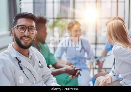 Ein junger Arzt mit Brille sieht in die Kamera vor dem Hintergrund des Diskus seiner Kollegen Stockfoto