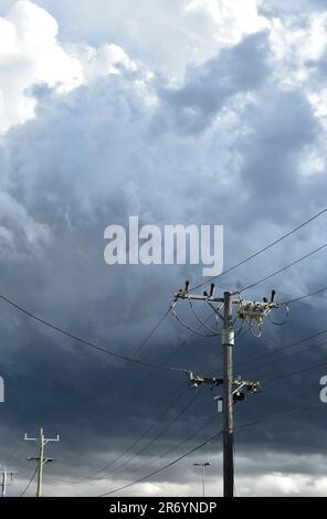 Ein vertikaler Schuss eines Strommastes, der Energie durch Transformatoren und Leitungen gegen einen stürmischen Himmel transportiert Stockfoto