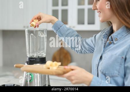 Wunderschöne junge Frau, die Banane in den Mixer gibt, um leckeren Smoothie in der Küche zu genießen Stockfoto