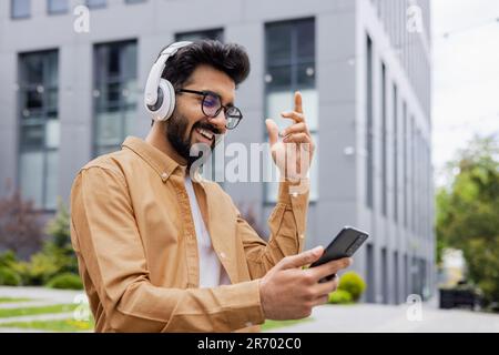 Ein junger, glücklicher Hindu-Mann, der auf der Straße mit Kopfhörern tanzt, ein Mann, der eine Online-App nutzt, um Musik zu hören und Radio zu hören, ein Student im Hemd, der durch die Stadt spaziert. Stockfoto