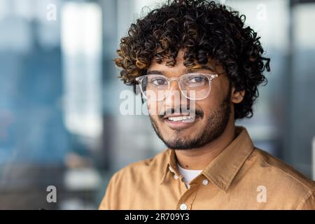 Nahaufnahme eines jungen hispanischen Mannes mit Brille, lächelndem Mann und Blick auf die Kamera am Arbeitsplatz im Büro, Programmierer zufrieden mit Erfolg und Erfolg. Stockfoto