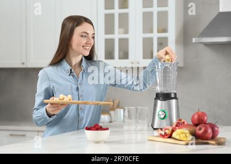 Wunderschöne junge Frau, die Banane in den Mixer gibt, um leckeren Smoothie in der Küche zu erhalten Stockfoto