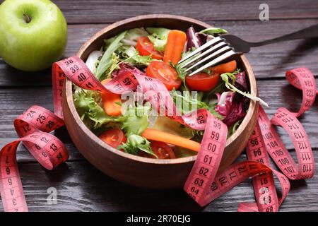 Schüssel mit frischem Gemüsesalat, Gabel, Apfel und Maßband auf Holztisch, Nahaufnahme. Gesundes Diätkonzept Stockfoto