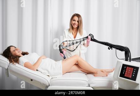 Porträt eines professionellen Kosmetikers, der gerne eine fraktionierte Laser-Hauterneuerung für eine lächelnde Frau durchführt, die mit innovativen Geräten arbeitet. Stockfoto