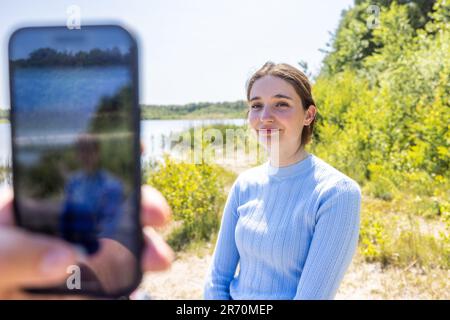 Lächelndes junges Paar, Familienfrau in blauer Kleidung, ein Mann fotografiert auf dem Handy seine hübsche Frau bei Sonnenuntergang im Freien an einem Seeufer am Sommertag. Hochwertiges Foto Stockfoto